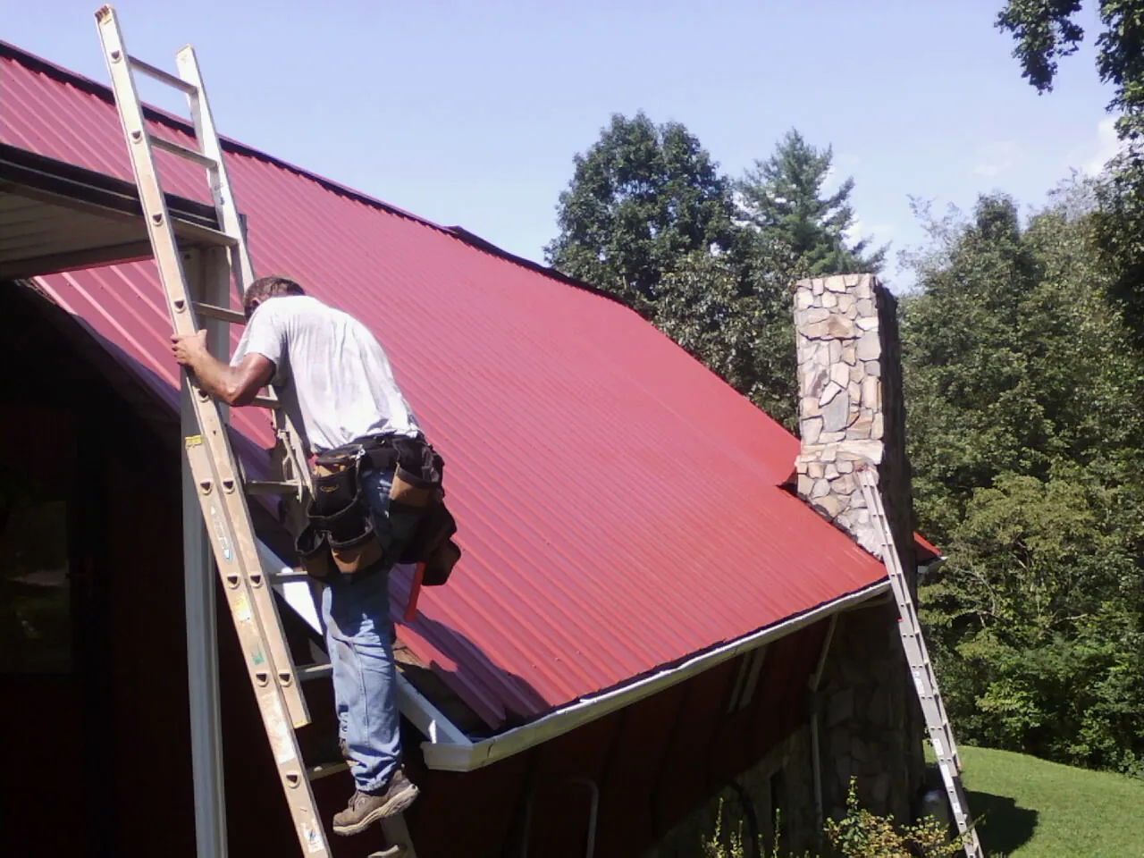 Roofer on a ladder beside a red metal roof and stone chimney — Soffit & Fascia Repair by Roofing Brothers in Kaibeto